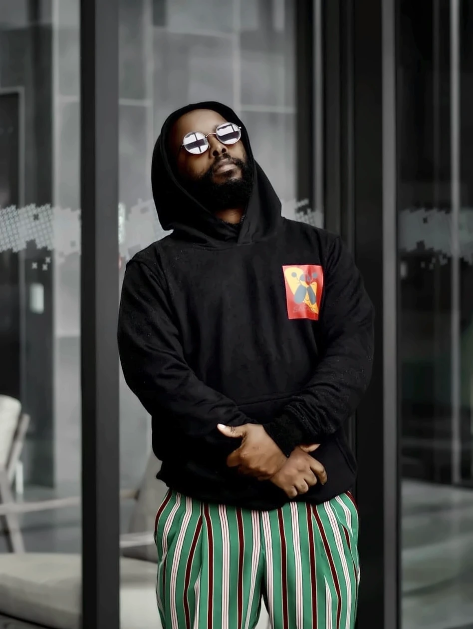 A man in a black SACXESS hoodie and striped trousers, head tilted back, standing against a glass wall